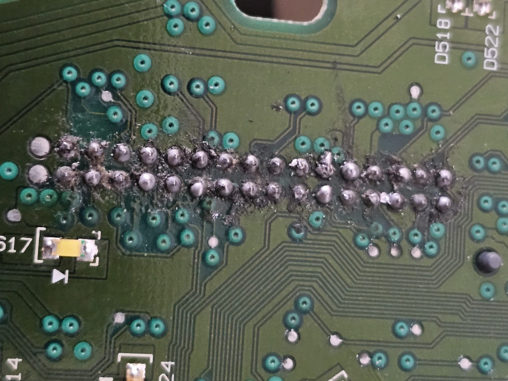 תקריב של לוח אם אלקטרוני (PCB) ירוק המציג שורת הלחמות לקויות ושרופות. נראים סימני חריכה קשים בגוון חום-שחור מסביב לנקודות החיבור של הרכיבים, המעידים על חימום יתר ושאריות פלקס שרוף.