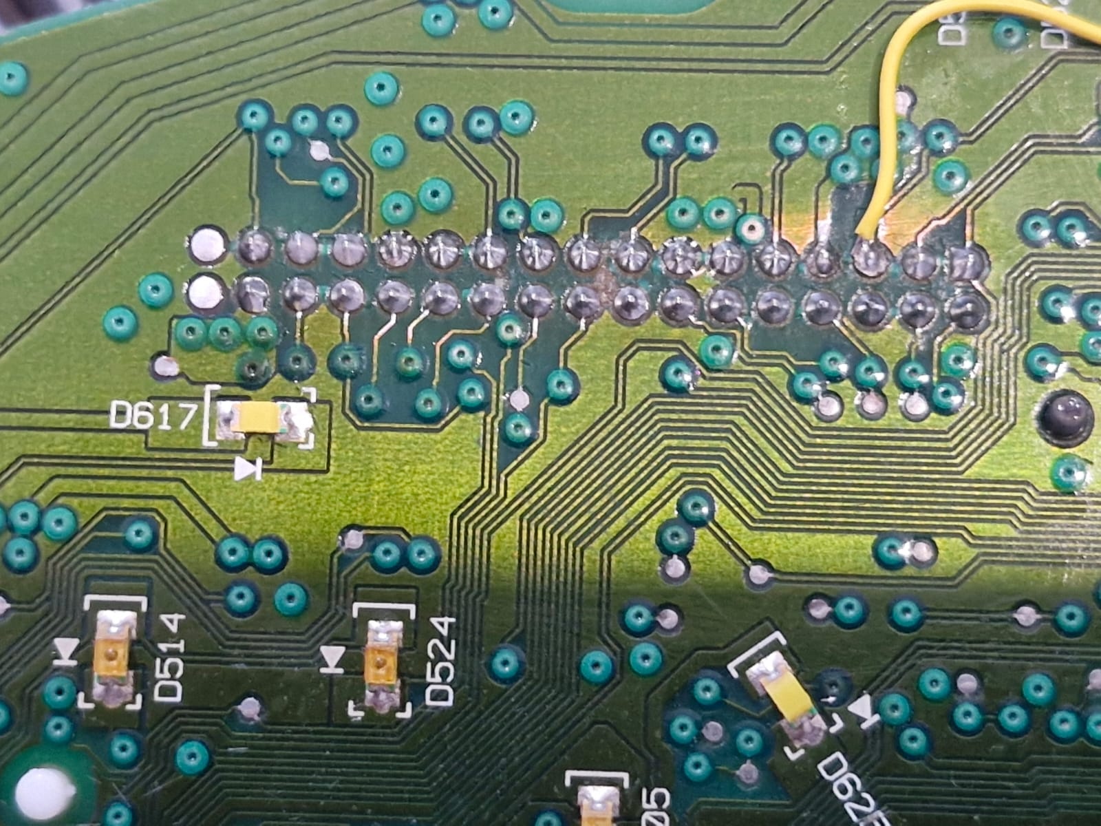 תקריב של לוח אם אלקטרוני (PCB) ירוק לאחר תיקון מקצועי ונקי במעבדה. נקודות ההלחמה מבריקות ותקינות, ונראה חוט ג'מפר (Jumper Wire) דק בצבע צהוב המולחם בצורה מדוייקת ונקי משאריות, כחלק משחזור מוליך בלוח.